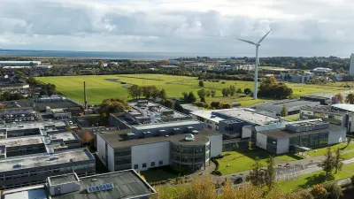 Aerial view of industrial buildings and farmland with wind turbines in the distance.