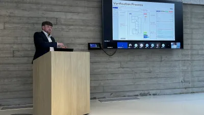 Person in business attire at wooden podium presenting with data displayed on large screen.