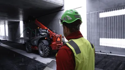 Construction worker in safety vest and green helmet examining a motorcycle in a parking garage.