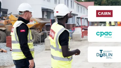 Two construction workers in safety vests and hard hats at a building site, with company logos visible.