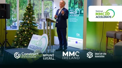 A person speaking at a podium with a Christmas tree and event sponsor banners in the background.