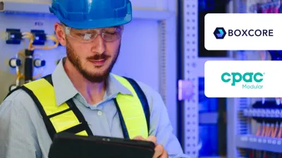 Worker in blue hard hat and safety vest using tablet near electrical control panel.