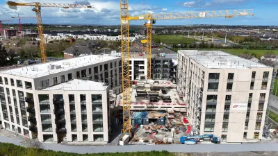 Aerial view of a large multi-story building under construction with yellow cranes and surrounding greenery.