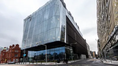 Modern glass and steel office building on an urban street corner, with a red brick building nearby.