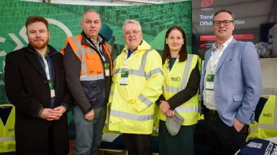 Five people posing together outdoors, two wearing high-visibility safety vests, at an event with green banners.