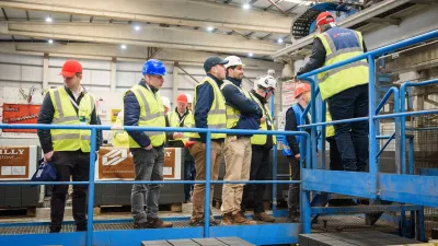 Group of people in hard hats and safety vests on an elevated platform inside an industrial facility.