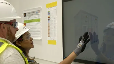 Two workers in hard hats and safety vests operating an industrial touchscreen panel.