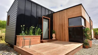 Small modern cabin with black and wood exterior, wooden deck, and large windows on a sunny day.