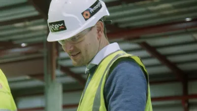 Man in white hard hat and yellow safety vest reviewing documents at a construction site.
