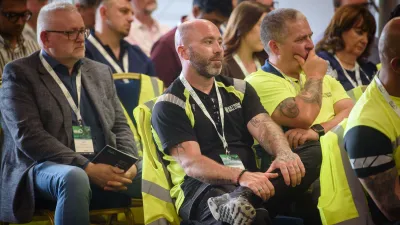 Audience members seated at an event, some wearing yellow safety vests and conference lanyards.