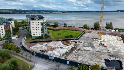 Aerial view of a construction site near a waterfront, with a crane, cleared land, and a white apartment building nearby.