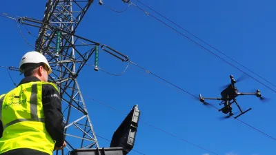 Worker in high-visibility vest inspecting power transmission tower while a drone flies nearby against a clear blue sky.