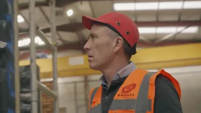 Man in orange safety vest and red hard hat looking sideways in an industrial warehouse.