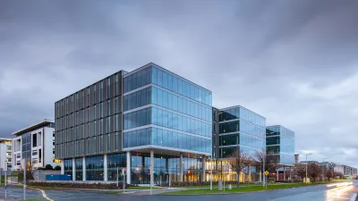 Modern glass office building with illuminated interior under a cloudy dusk sky, urban setting.