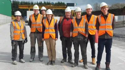 Seven people in orange safety vests and white hard hats posing outdoors at a construction site.