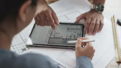 Two people reviewing architectural floor plans on a tablet, with printed blueprints on the desk.