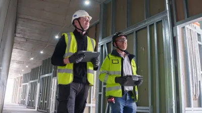Two construction workers wearing hard hats and high-visibility clothing, inspecting a building interior while holding tablets.