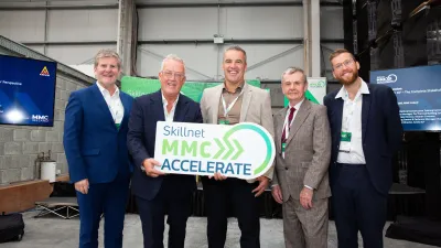 Five men in business attire smiling, holding a green "Skilletnet MCC Accelerate" sign indoors.
