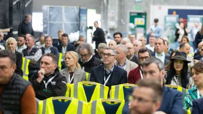 Large crowd of attendees at an indoor conference or trade show, some wearing yellow safety vests.