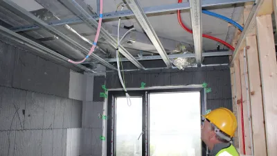 Construction worker in yellow hard hat inspecting ceiling with exposed pipes and wiring in an unfinished building.