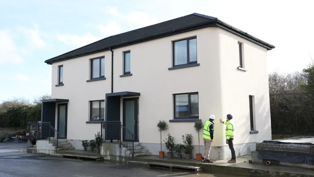 Two people in high vis jackets and hard hats inspect modern white semi-detached houses.