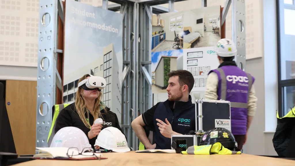 Two people sitting at a table, with the male watching as the female uses a VR headset.
