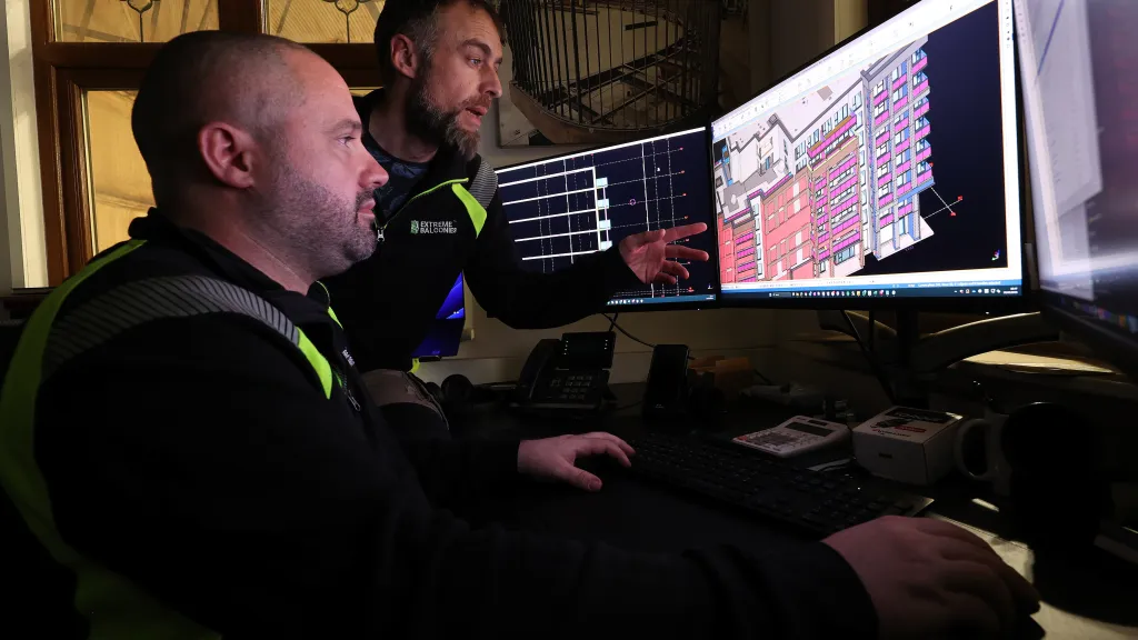A male points out something in a digital building design on a computer screen to another male.