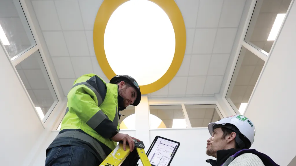 A male in a hard hat with a tablet shows the screen to another male in a hard hat standing on a ladder. 