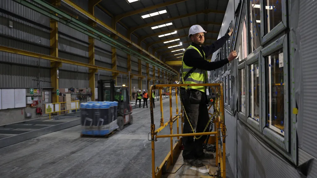 A male with a high vis vest and hard hat stands on a scissor lift to fit a window.