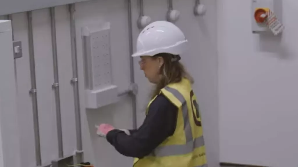 Electrician in high-vis vest and hard hat working on equipment near a control panel.
