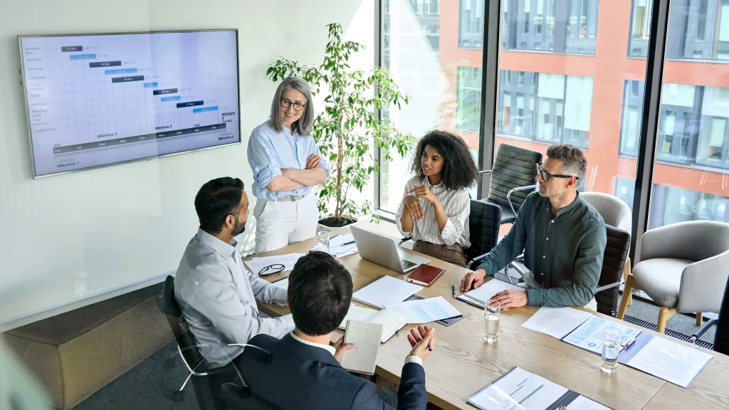 A group of people in business clothing with laptops and pieces of paper in front of them sit around a long table, talking.