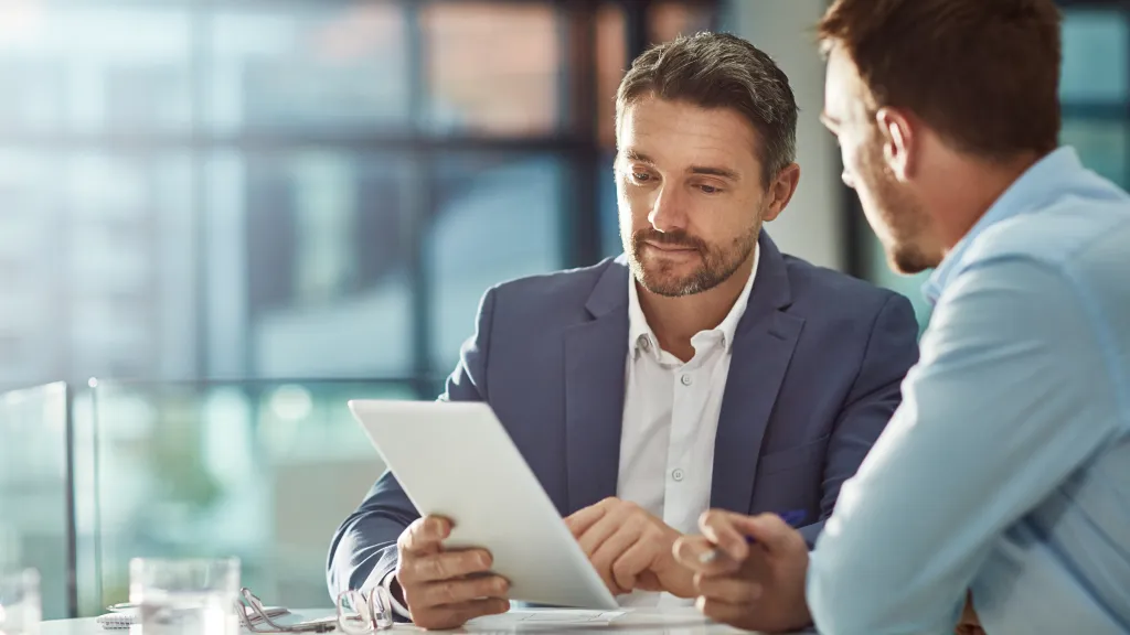 A male in a blue business suit reads from a tablet to another male in a business shirt.