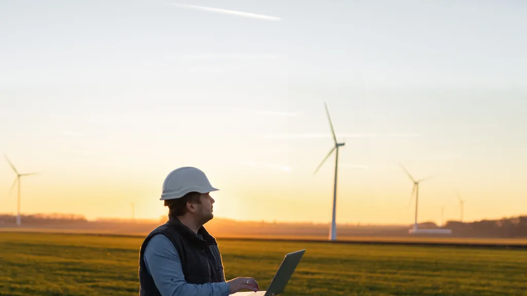 A male in hard hat and vest with an open laptop stands in a field with wind turbines in the background.