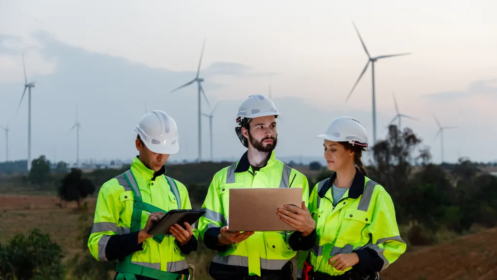 A group of people in high vis jackets and hard hats read from tablets standing in a field with wind turbines.