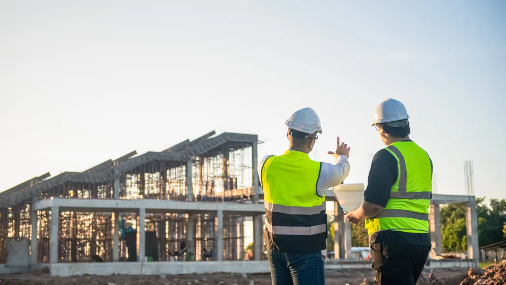 Two people in high-vis vests and hard hats standing at a construction site, looking at a building under development.