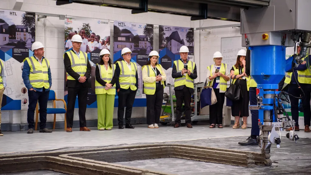 A group of people wearing high-visibility vests and hard hats standing in a row inside an industrial or construction facility, with machinery visible in the foreground.