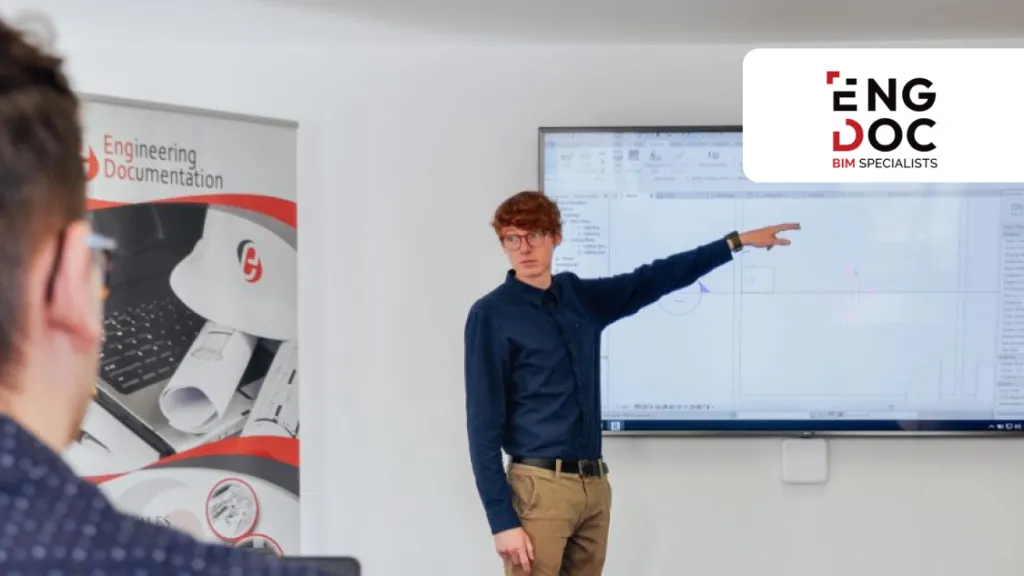 A person is standing next to a screen, pointing at a slide during a training session in a classroom.