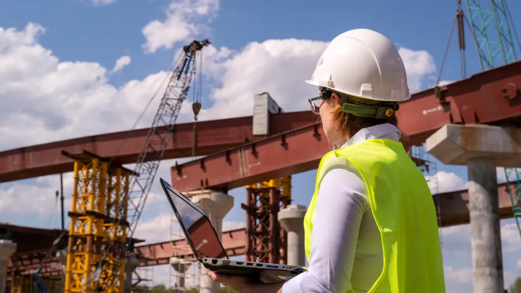 A construction worker wearing a high-visibility vest stands in front of steel structures.