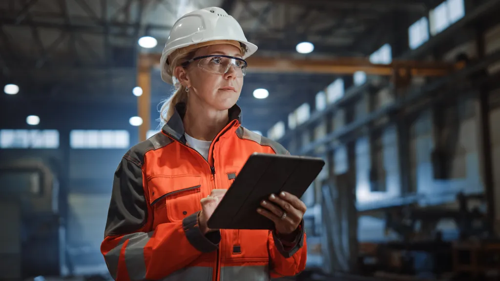 safety officer wearing a hard hat, safety glasses, and a high-visibility jacket, holding a tablet in an industrial warehouse.