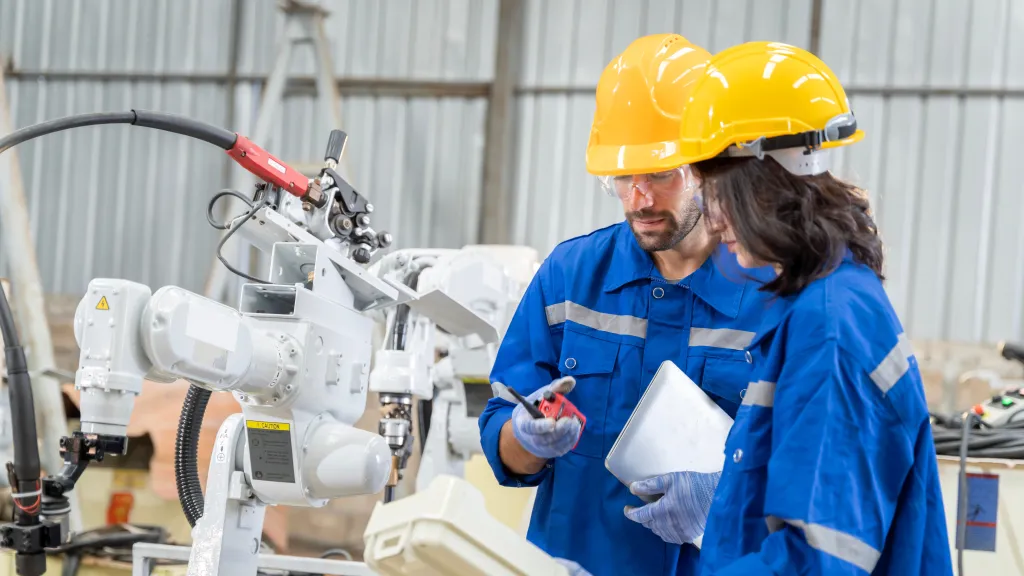 Two people wearing work clothes and yellow hard hats are standing next to a robot.