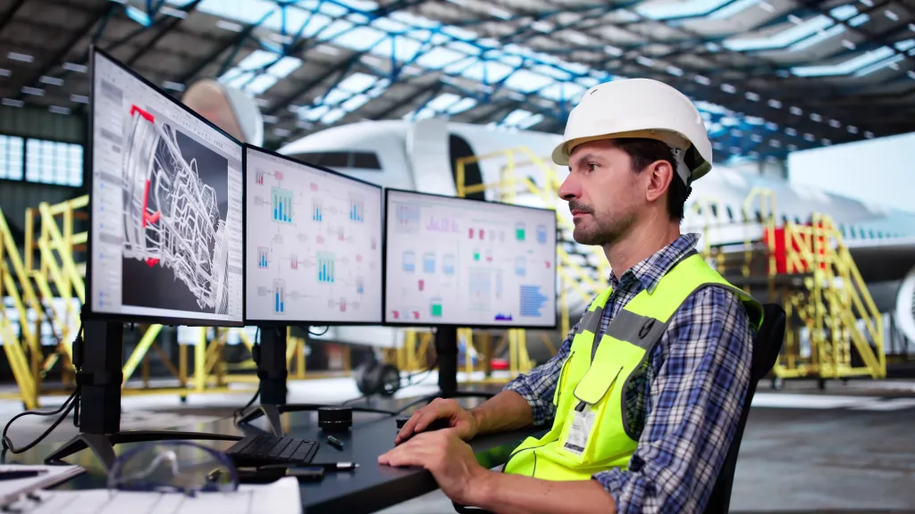 Man in safety vest and helmet monitoring construction plans on multiple computer screens.