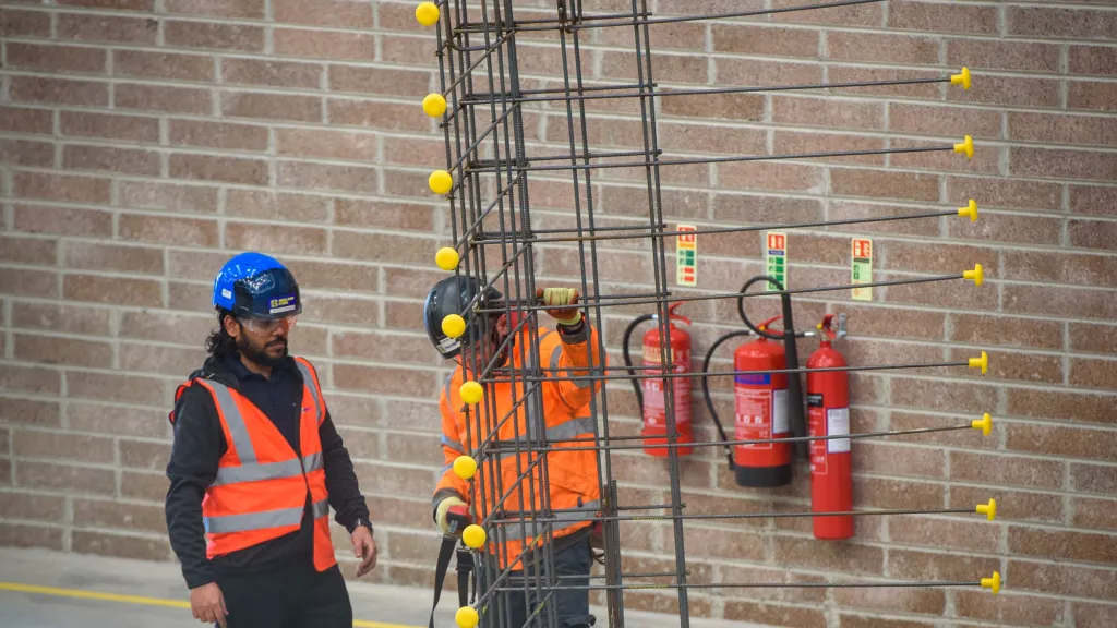 Two construction workers in safety gear standing near a vertical rebar structure inside a building under construction.