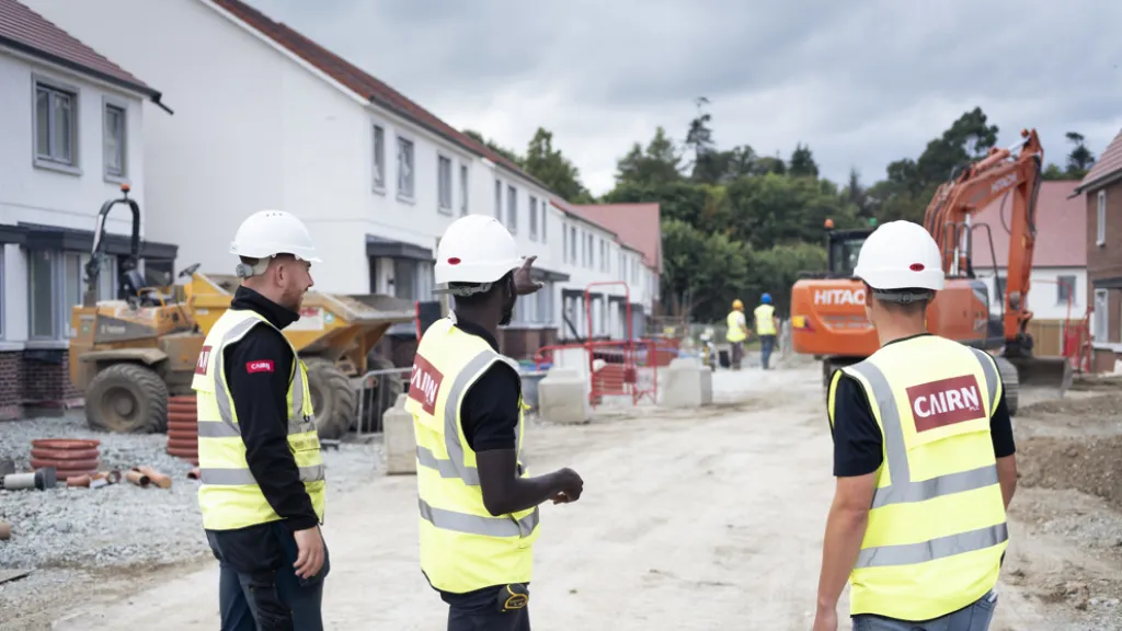 Three construction professionals in safety gear and hard hats are standing on a residential building site, discussing progress. Houses under construction are visible in the background.