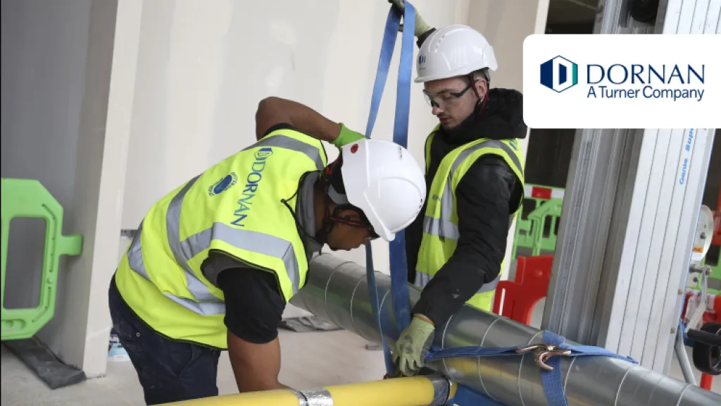 Two construction workers in high vis jackets, white hard hats, and goggles work on tying a pipe