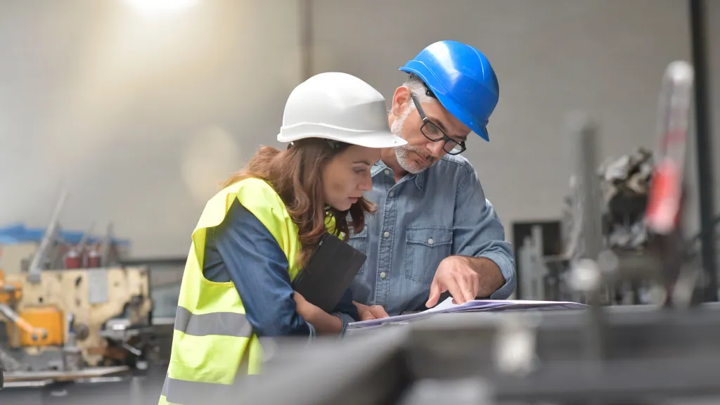 Two people wearing hard hats and safety vests reviewing a technical drawing in an industrial setting.