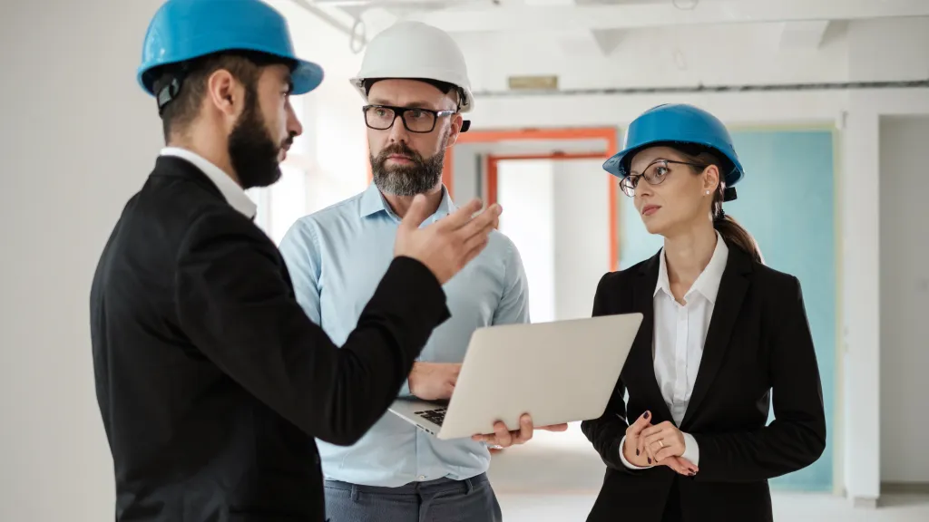 Three engineers wearing formal clothing and safety helmets discussing a construction project indoors, with one holding a laptop.