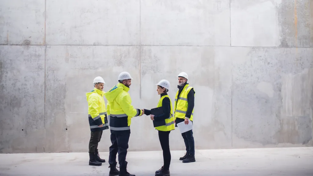 Four construction workers wearing high-visibility jackets and helmets standing and talking near a large concrete wall.
