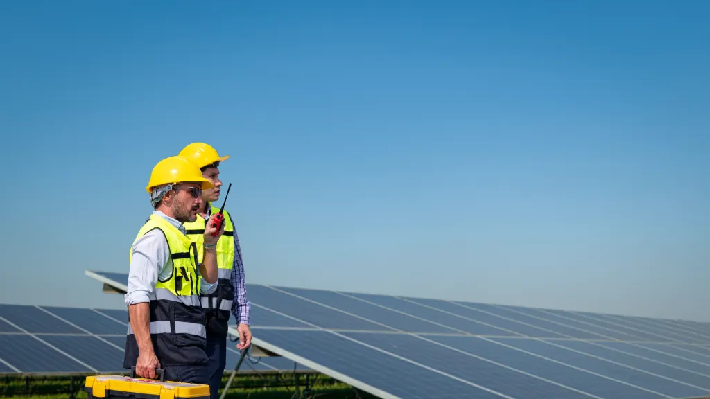 A construction worker wearing a yellow hard hat and safety vest stands beside a solar panel array, holding a toolbox in a sunny outdoor environment.