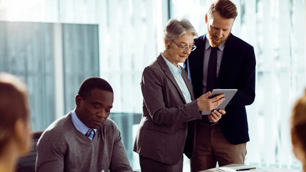 Group of professionals in a modern office having a discussion around a table, with one person showing a tablet to a colleague.