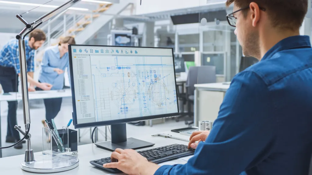 Person working at a desk with a computer monitor displaying technical engineering drawings,